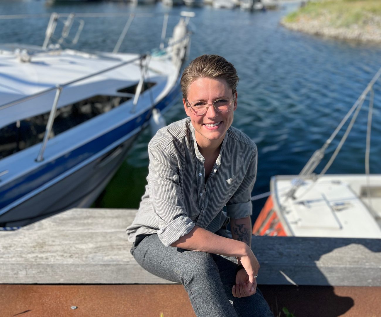 Sillan sidder smilende på kajkanten i havnen med sejlbåde i baggrunden.